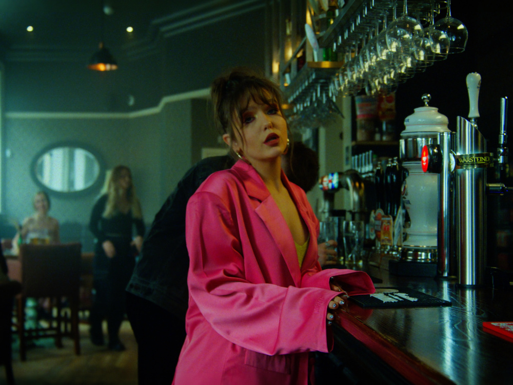 Still image from "The Weekend" music video which sees Faye Fantarrow at the bar, wearing a hot pink blazer and lime-green bra.