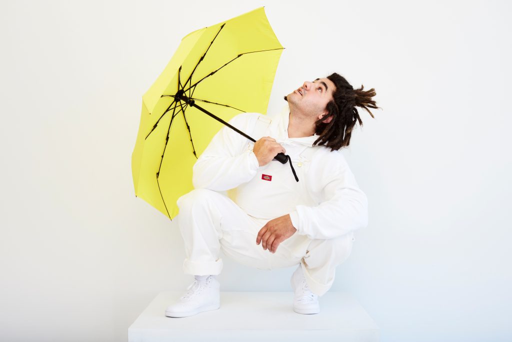 Promotional photo for "Summer Sunshine" which sees Fran Roz on a bright white photo set, wearing all white, with his brown hair tied up in a bun, and he's holding a yellow umbrella. He is crouched with the umbrella open and over his arm.