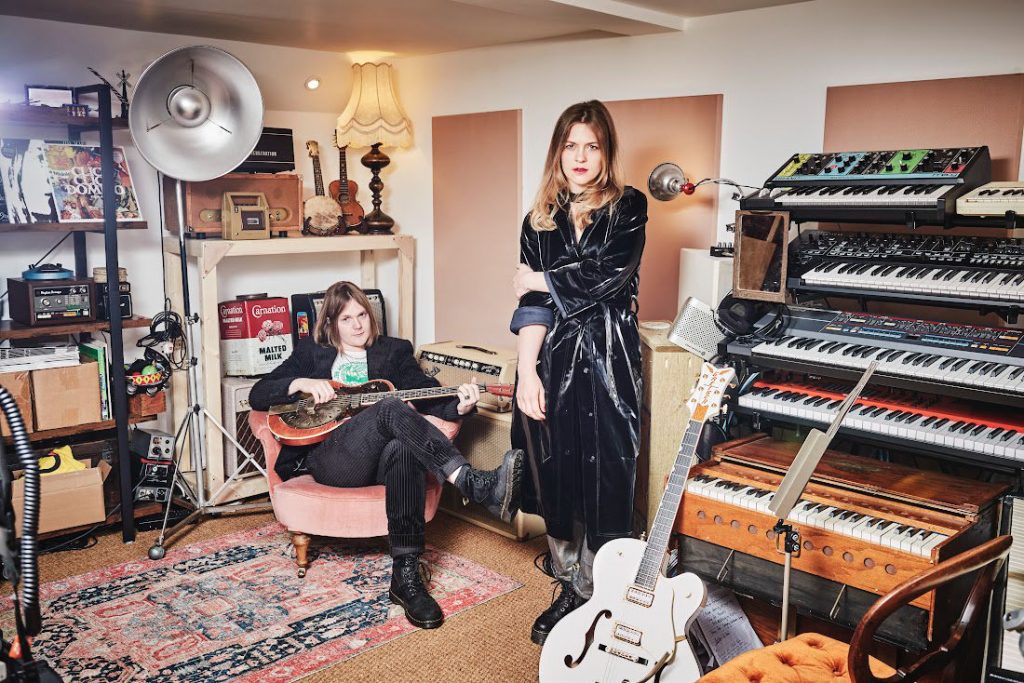 Promotional photo for "My Whispers Are Wildfire" which sees Ida Mae posing in a home studio full of keyboards, guitars, lights, and microphones, with a classic rug on the hardwood floor. Stephanie Jean Ward is standing wearing a black trench coat, while Chris Turpin is sat on a light pink chair with a guitar across his lap.