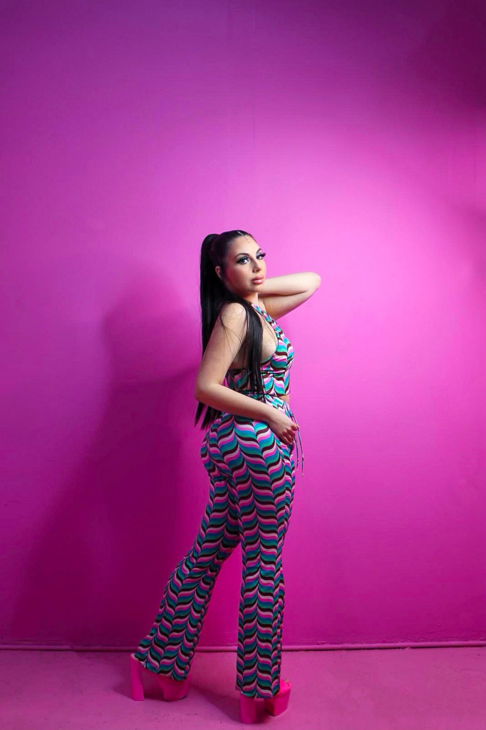 Promotional image for "My Hair" which sees Mandia posing in front of a pink background, looking over one shoulder at the camera, wearing a light blue jump-suit with pink and cream lines patterned over it. Her brown hair is tied back in a ponytail, and she's wearing bright pink platform heels,