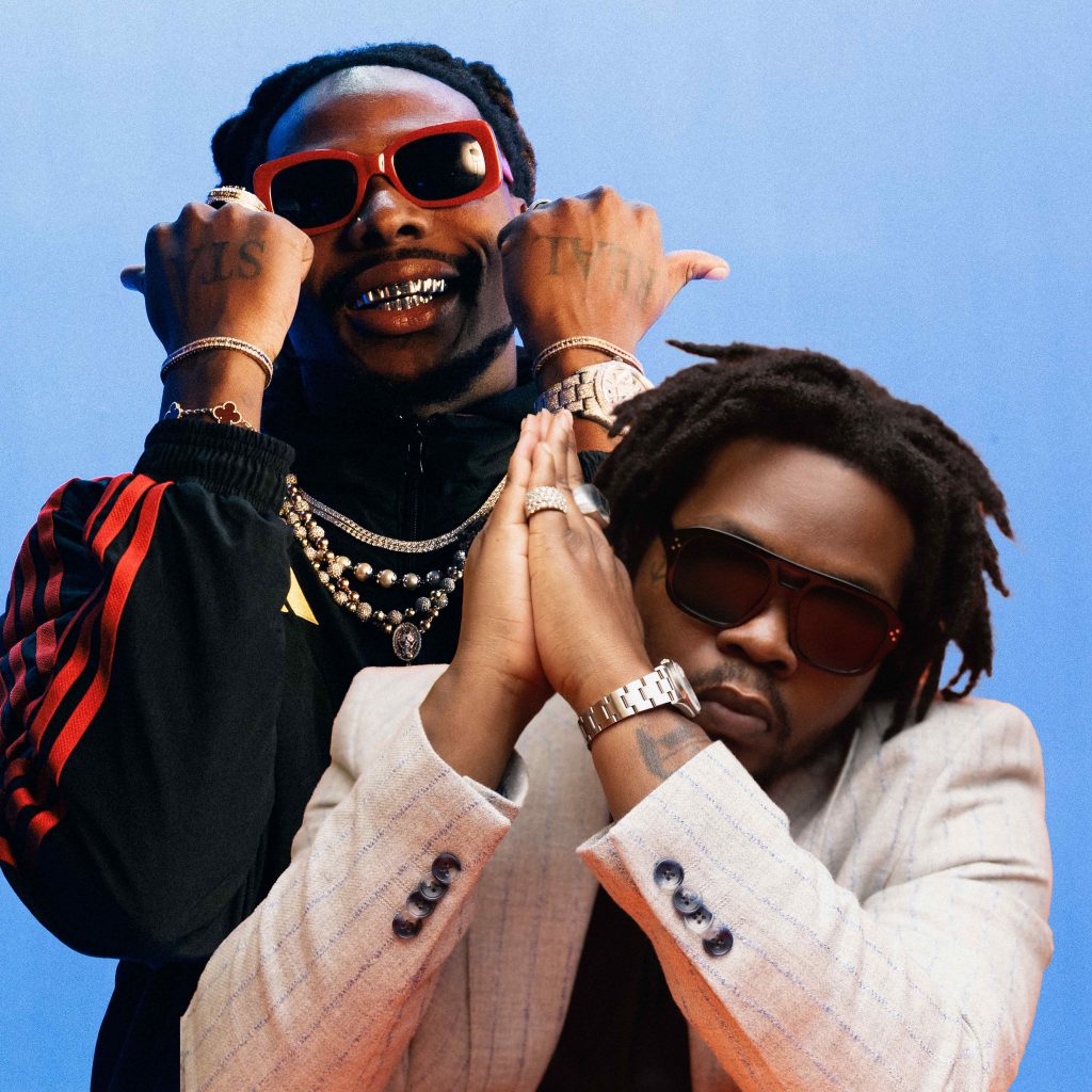 Promotional photo for "New Religion" which sees Olamide and Asake together against a blue background. Olamide is sitting down with his hands up to his head in a prayer sign, he is wearing a white jacket and dark shades. Asake is behind him with his arms up to his head with his hands in fists, showing off his bracelets. He is wearing a black jacket with red sleeve design, a few silver chain necklaces and some red-rimmed sunglasses, and he is grinning.