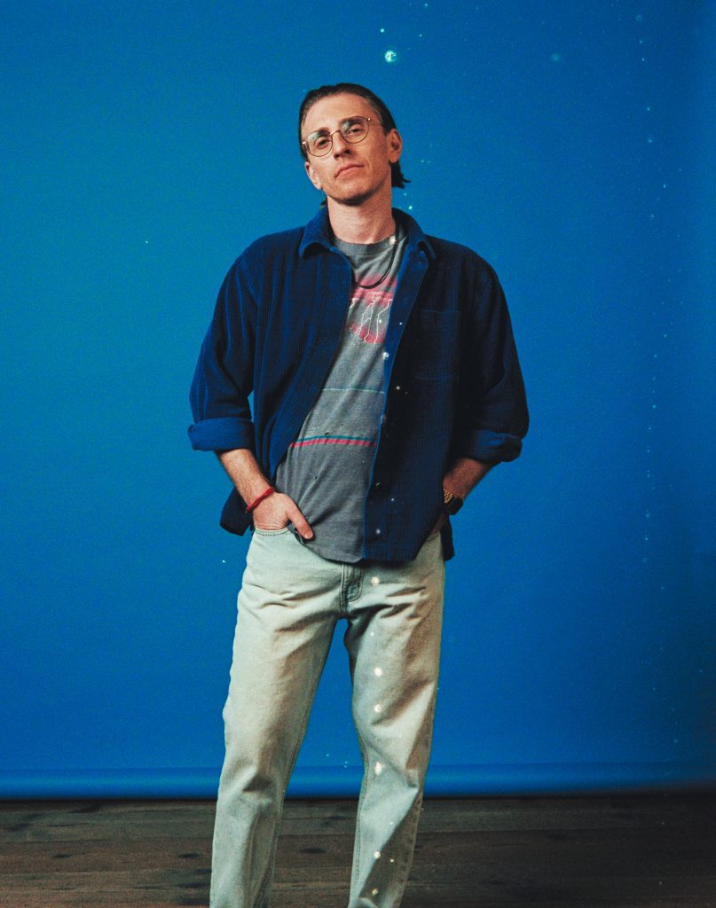 Promotional photo for "retrospection (19:30)" which sees Payson Lewis standing in front of a blue background, with his hands in his light-blue jean pockets. He is wearing a dark blue long-sleeve shirt opened over a grey t-shirt.