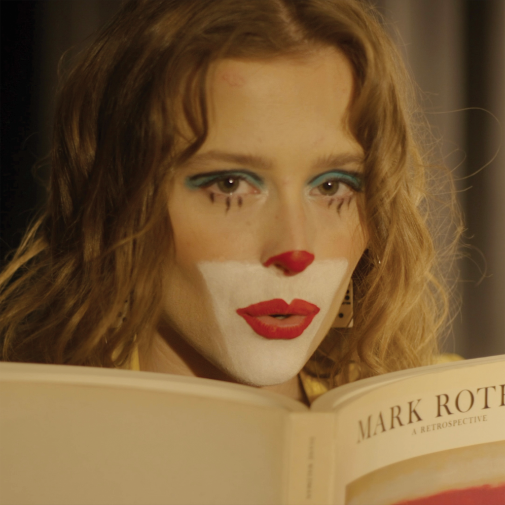 Promotional photo for "Ride or Die" which sees a head-shot of Puma June with her clown make-up but pretty, with a white lower-face, red lipstick, a red nose, blue eye-shadow and drawn-on eyelashes. She's holding an open book up to her chin, and her blonde-brown hair cascades as waves down either side of her face.