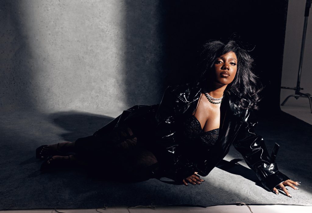 Promotional image for "So Fine" featuring Duduboy, which sees Rukmani in a concrete setting, lying on her side with her left hand on the ground with her arm locked for support. She is wearing a black leather jacket over a mesh outfit over a black glittery bra. She's wearing two chain necklaces and her black shoulder-length hair is slightly windswept