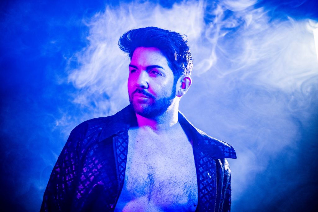 Promotional photo for the pride edition series of Celebrity Skin Talk which sees Scott McGlynn surrounded by smoke, with his black shirt open, revealing his chest. There's a bright blue colour filter to the image.