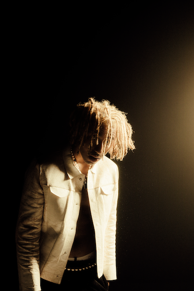 Promotional photo for "sodium lights" which sees Xadi surrounded by darkness, with a white spotlight hitting him from off-camera, showcasing his cream denim jacket, brown mid-length afro hair and the black t-shirt he's wearing under his jacket.