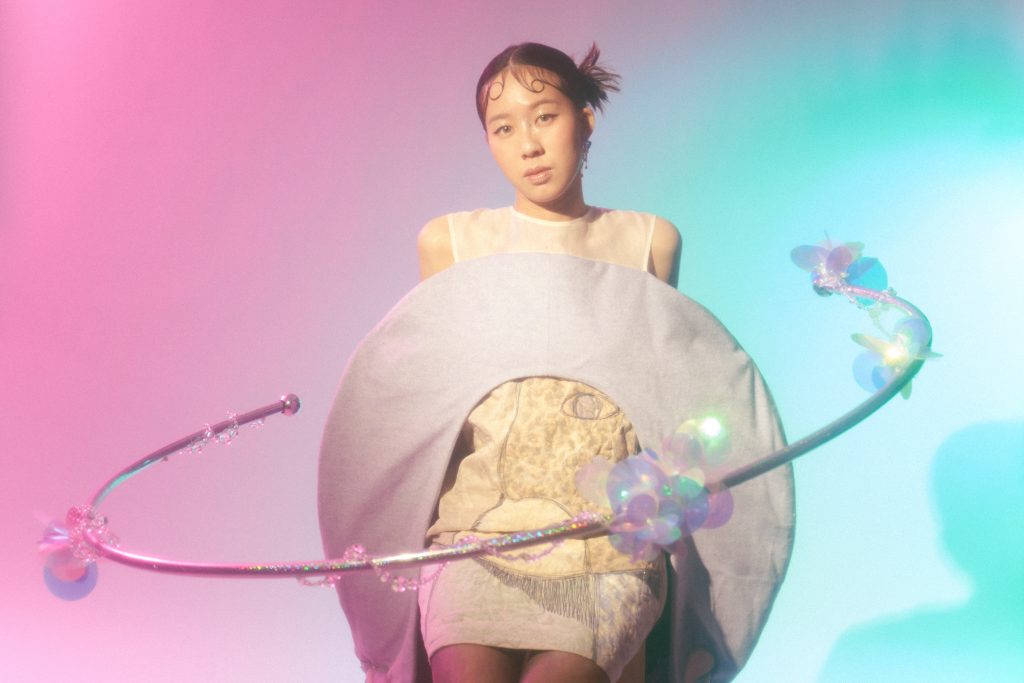 Promotional photo for "APOLLO-13" which sees XTIE in a cream dress in front of a pink to blue ombre wall, with two grey circle hoops around her with water splashing filter effects.