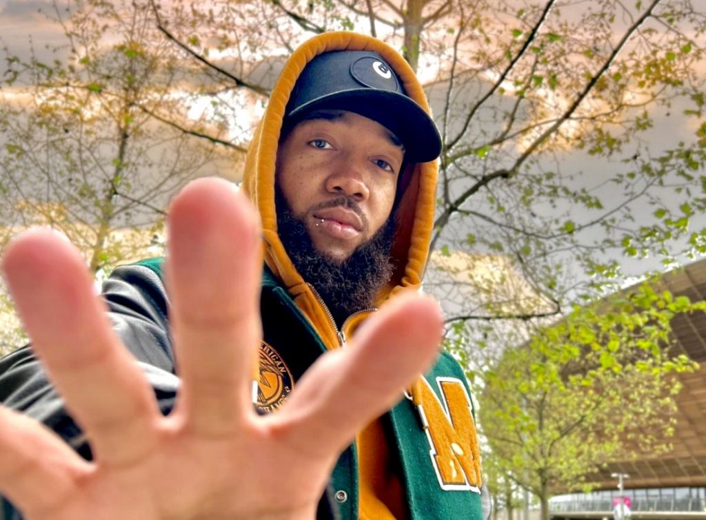 Promotional photo for "The Blckout" which sees XVR BLCK looking at the camera with his hand reaching for the lens. He is earing a black cap under a yellow hoodie, with his beard growing prominently out the hoodie. He is also wearing a green varsity jacket and there is a tree behind him.