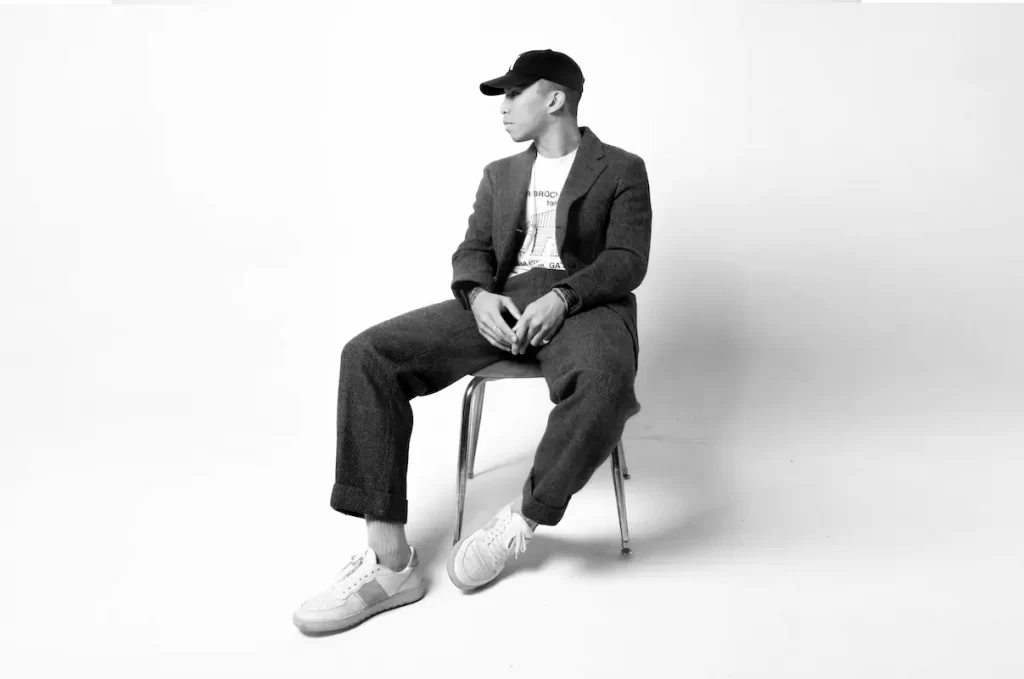 Promotional photo for "One Sided" featuring Abby Holliday, which sees Yueku sitting on a chair on a photo set. wearing a suit with trainers and a snapback cap, and he is looking towards the left. The photo is in black and white.