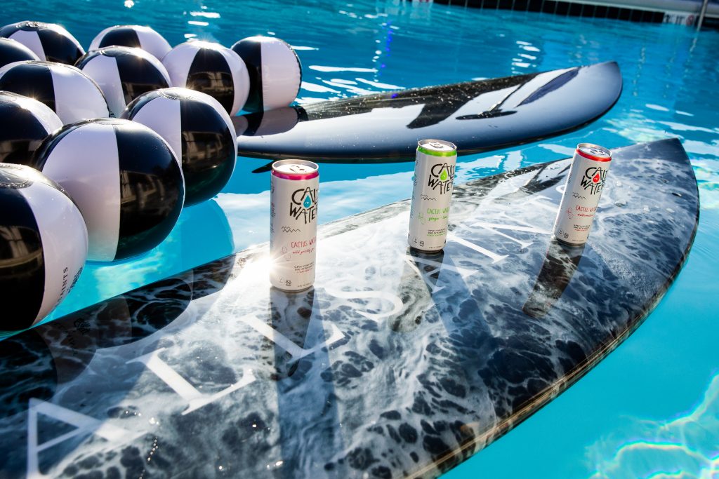 Promotional photo for AllSaints and Caliwater Sunglasses rooftop event. The photo sees two black surfboards with the AllSaints name on them, with black and white beach balls to the left, and the three Caliwater flavoured cans on the nearest board.