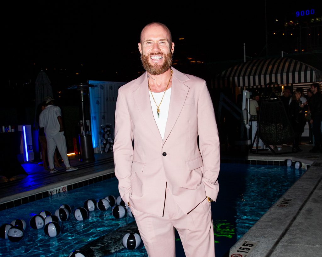 Oliver Trevena posing in front of a swimming pool, at night, wearing a pale pink linen suit jacket and trousers with a white t-shirt underneath. He has his hands in his trouser pockets and he is smiling.