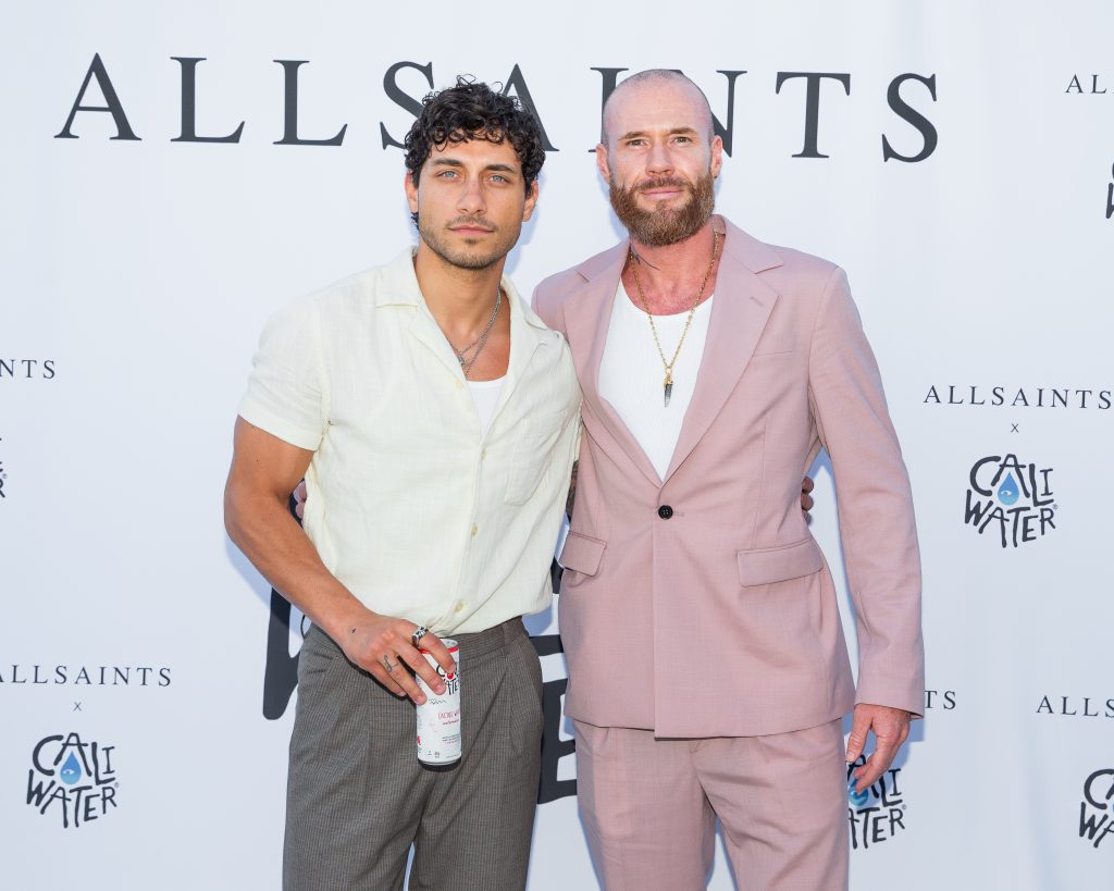 Rob Raco posing with Oliver Trevena in front of the AllSaints x Caliwater Summer Sunglasses Event backdrop with Rob Raco wearing grey trousers and a white shirt, and Oliver Trevena wearing a pale pink linen suit.