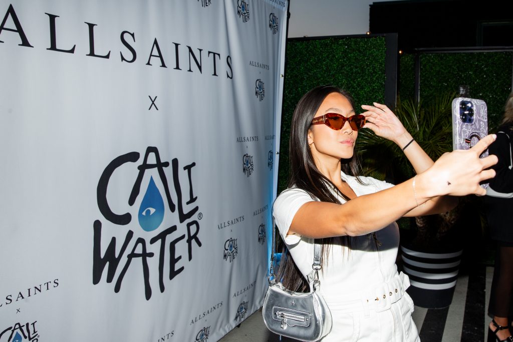 Tammy Kay Ly taking a selfie with the AllSaints Summer Sunglasses eyewear, whilst wearing a white dress, and with the AllSaints x Caliwater sponsor backdrop behind her.