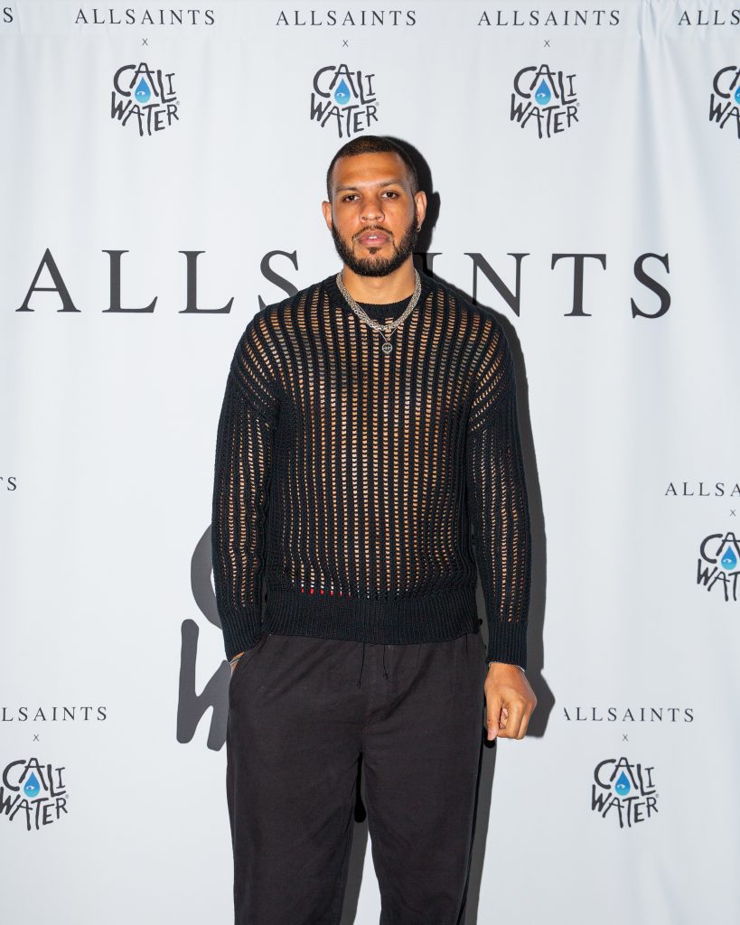 Sarunas J. Jackson posing in front of the AllSaints x Caliwater Summer Sunglasses Event backdrop, wearing a striped-mesh black sweater and black trousers.