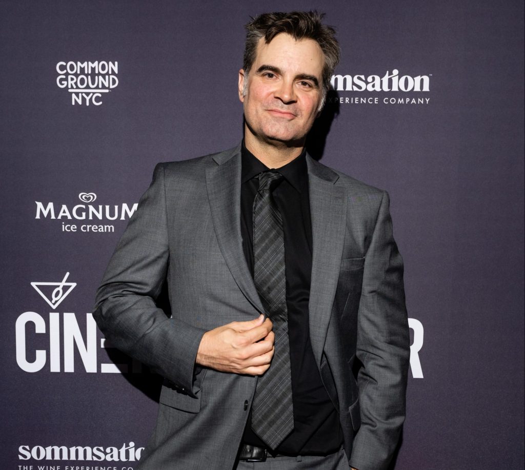 Joe Lynch posing in front of a sponsor backdrop at his film premiere events, in a grey suit.