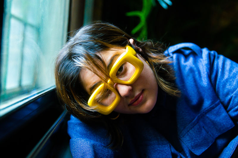 Promotional photo for "V.I.P" which sees Belot leaning against a window sill, tilting her head right over so her brown hair falls down her side and around her yellow block glasses and her blue shirt.