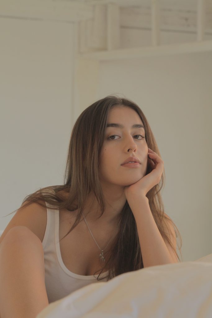 Promotional photo for "Linen" which sees Ella Rossi wearing a white vest top, sitting on the floor with one elbow resting on her bed with her hand under her chin, almost in a bored expression.