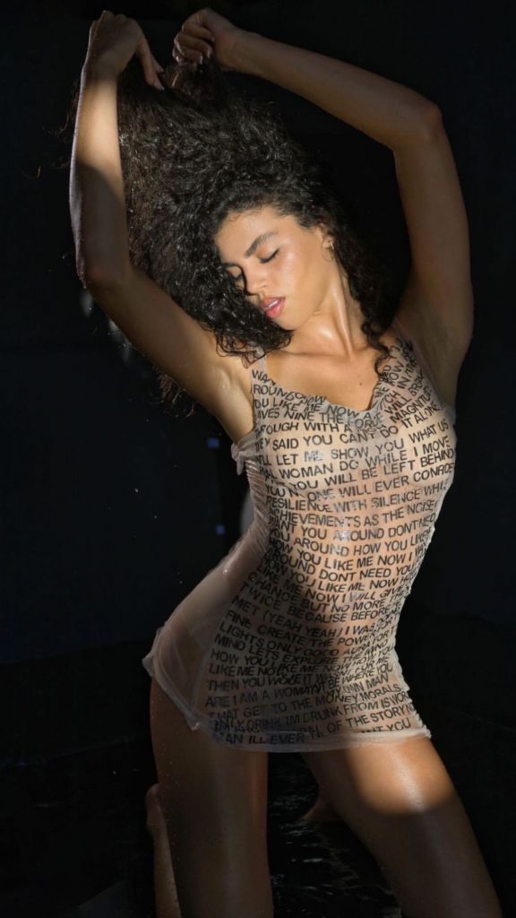 Promotional photo for "Mueve Tu Cuerpo" which sees Emika Love posing with her arms in the air, loving life, with a see-through beige dress with words patterned across the front saying positive things about being a woman. Her curly brown hair is pushed up and away from her beautiful face.