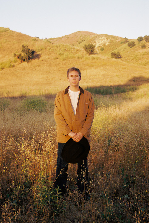 Promotional photo for "Got Him!" which sees Ethan Tasch standing in a wheat field wearing a white t-shirt under a wheat-coloured jacket and blue jeans. He is holding a black cowboy hat in his hands.