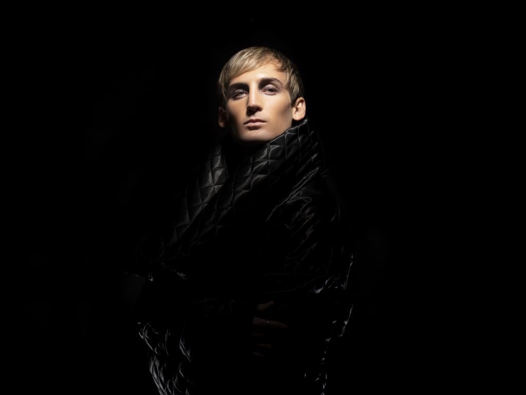Promotional photo for "3 Days, 2 Nights" which sees PLS&TY posing in a black quilted plastic coat which he is wrapping around himself as he looks at the camera while facing to the left. The coat melds into the black background while his face is spotlighted, with his blonde hair and pale skin having an angelic glow.