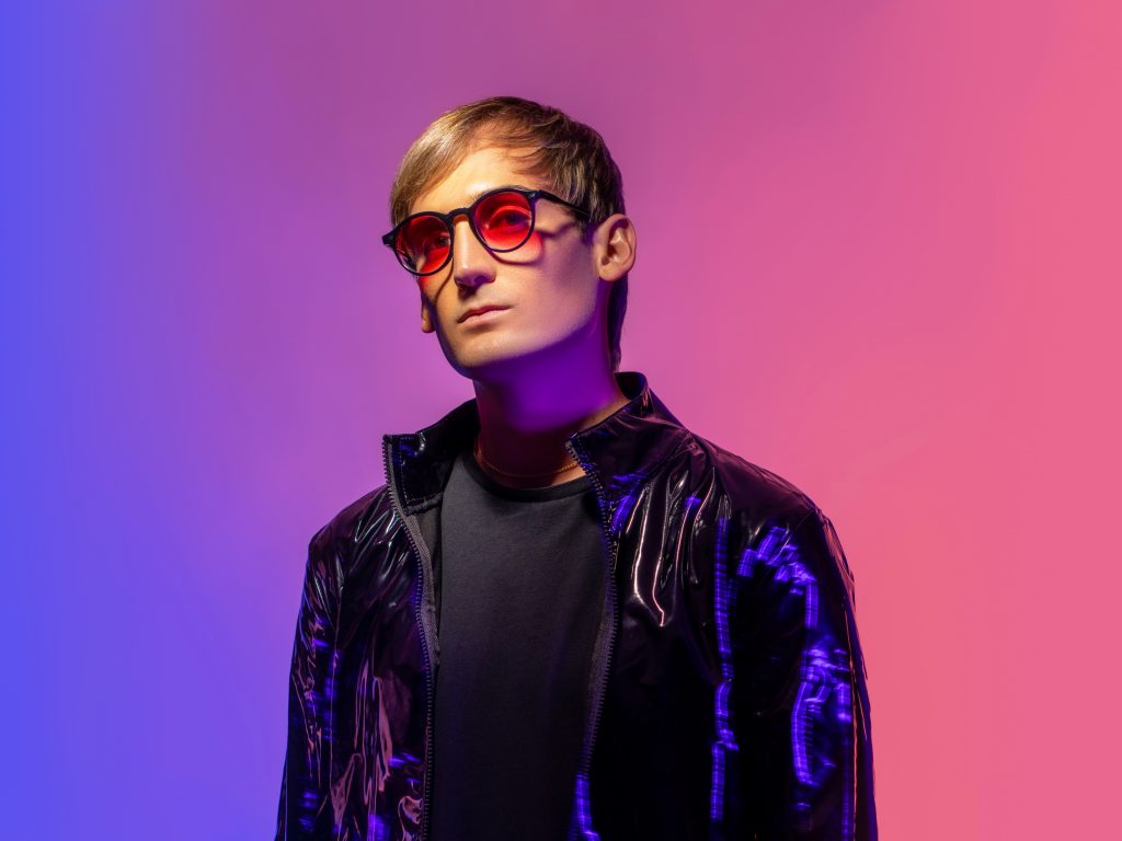 Promotional photo for "3 Days, 2 Nights" which sees PLS&TY standing in front of a pink-to-purple ombre background from right-to-left. He is wearing orange shades, a black biker jacket over a black t-shirt.