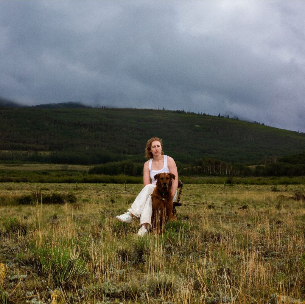 Promotional photo for "Diamond Ring" which sees Hallie Spoor sitting on a wooden chair in a grass field with a brown dog sitting in front of her. She's dressed in a white top and white trousers and the sky is cloudy.