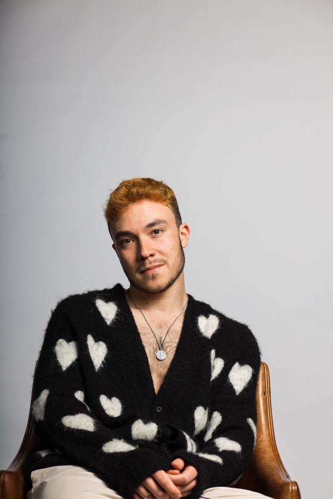 Promotional photo for "Glimpses" which sees Fox Jackson sitting on a wooden chair, wearing a black cardigan with white hearts all over it, with a low neckline, showing off his chest and silver necklace.