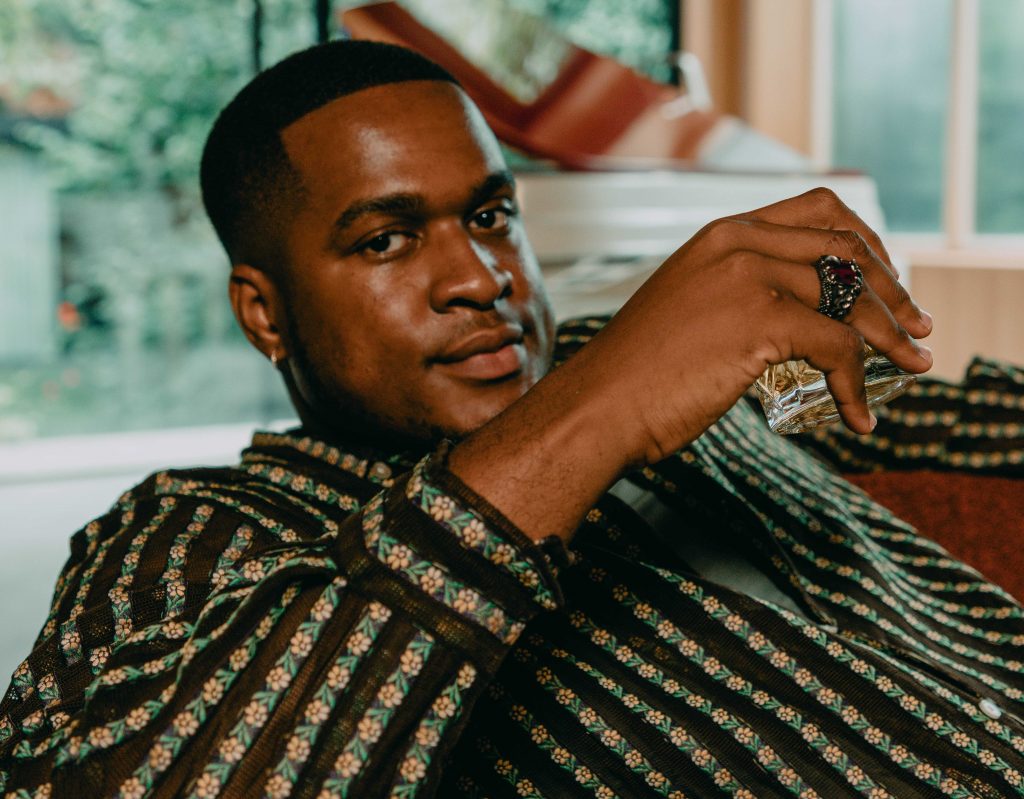 Promotional photo for "Hear Me Out" which sees Gabzy sitting on a red couch looking at the camera with his arm out in front of him. He is wearing a pale-green and brown checkered with flowers shirt and a huge black ring on his finger.