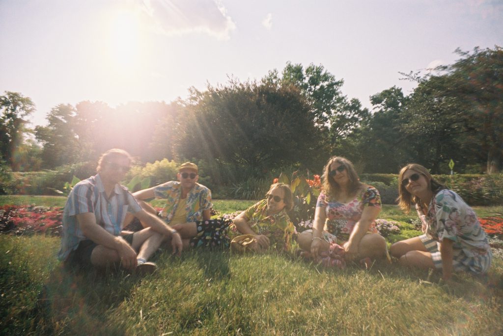 Promotional photo for "Boyfriend" which sees the band members of Hot Freaks sitting in a semi-circle at a park, on the grass.