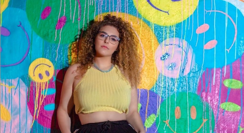 Promotional image for "Compare to Me" featuring Jamie Love, which sees Kapture leaning against a painted wall with different brightly-coloured smiley faces on it. She's wearing a yellow sleeveless top and black trousers. Her light brown afro hair reaches just past her shoulders and she completes her look with purple-rimmed glasses and a silver chain necklace.