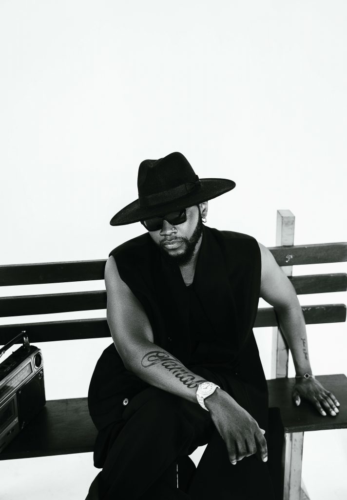 A black-and-white promotional photo for "Maverick" which sees Kizz Daniels sitting on a bench wearing a wearing a gilet jumper, with a broad-brimmed hat and dark shades, with one arm across his legs showing off his tattoo that says "Jamal" in cursive text all along his forearm.