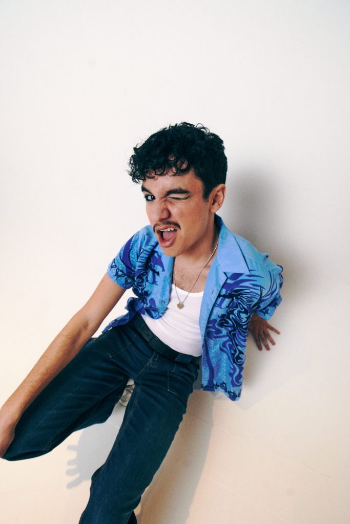 Promotional photo for "Don't Call Me At A Party" Which sees Liam Benayon wearing a white vest top under an open blue shirt with dark blue palm tree design on the sides, and some dark blue jeans. He's winking at the camera.