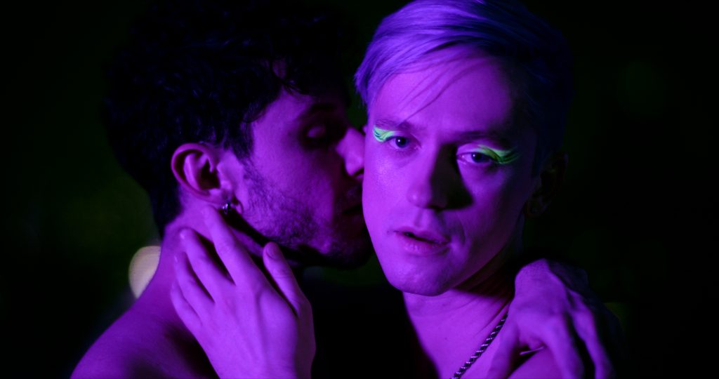 A still from the "Until Tomorrow" music video which sees Hernando Umana kissing the cheek of Magnus Riise as the singer looks at the camera with a lustful gaze. They're both shirtless, Magnus Riise's hand is on Hernando Umana's neck and the singer has luminous green eye shadow on.