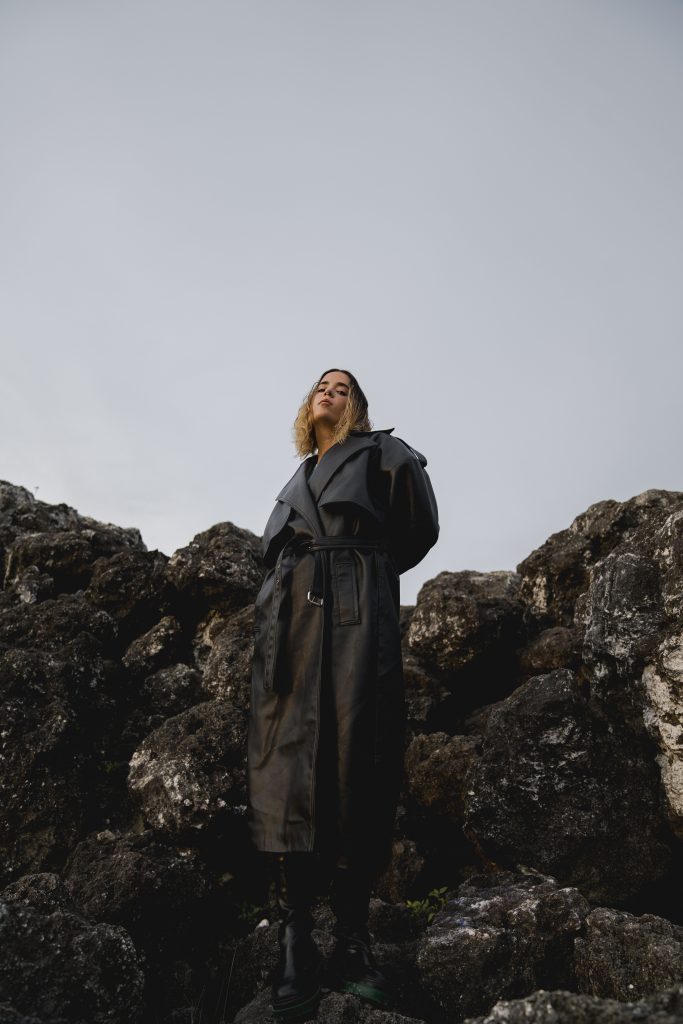 Promotional photo for "sometimes a person never comes back (but that's okay)" which sees MAVICA on a huge rock formation wearing a green trench coat and posing to the side.