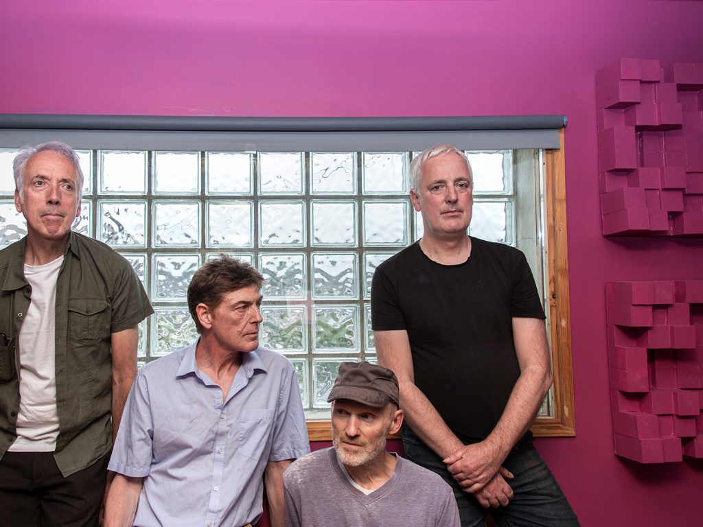 Promotional photo for "The Good, The Real, The Beautiful" and "The Strange Rain" double single drop which sees the band Otherish standing against a window that has purple-pink walls. There are four of them and are all looking in different directions.