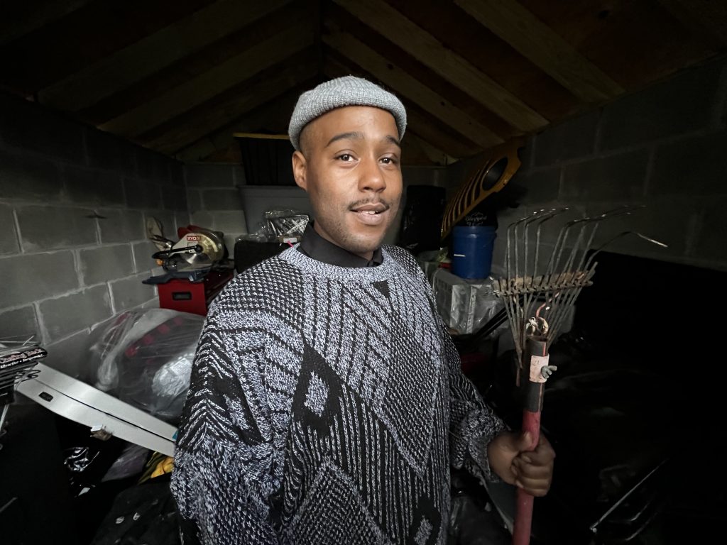 Promotional photo for "Betcha Can't Do It Like Me" which sees Ray Hodge in a grey sweater and a grey beanie, holding a rake while standing in a garage.