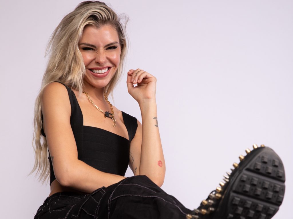 Promotional photo for "Munca" that sees Sam Blacky posing at a photo studio, sitting down against a white background. She's wearing a black halter top with black cargo trousers and black boots, and she's laughing as she leans backwards.