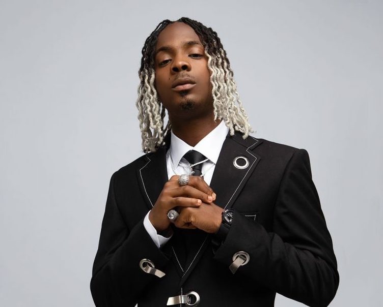 Promotional photo for "Sharpally" which sees Young Jonn posing in a photo studio, in front of a grey background, wearing a dark black suit with a white shirt and a black tie, with his dreadlock hair two-tone with blonde half-way while the rest is black to his scalp. His hair is shoulder length and parted down the middle. He is holding his hands together out in front of his chest.
