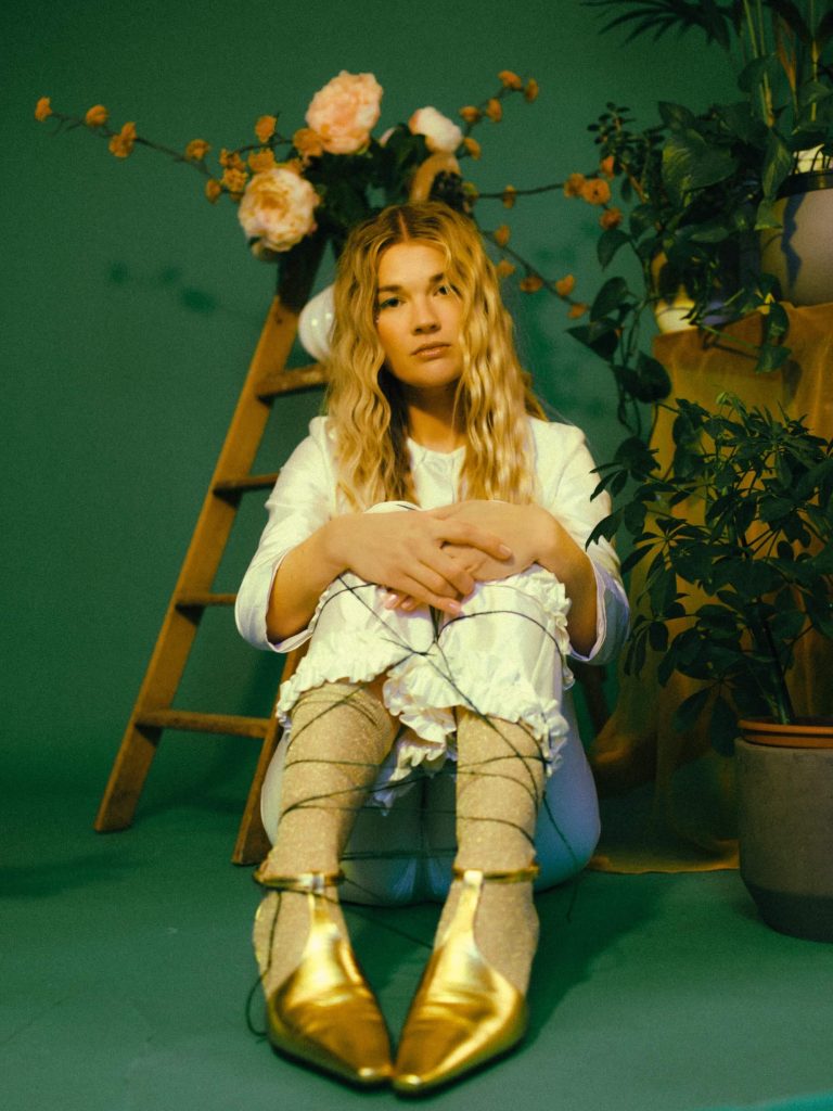 Promotional photo for "Where I Go" which sees PEARL with her knees to her chest and her arms around her legs, wearing a white shirt and matching trousers with gold boots that match her wavy blonde hair. She's sitting in a royal green-painted room.