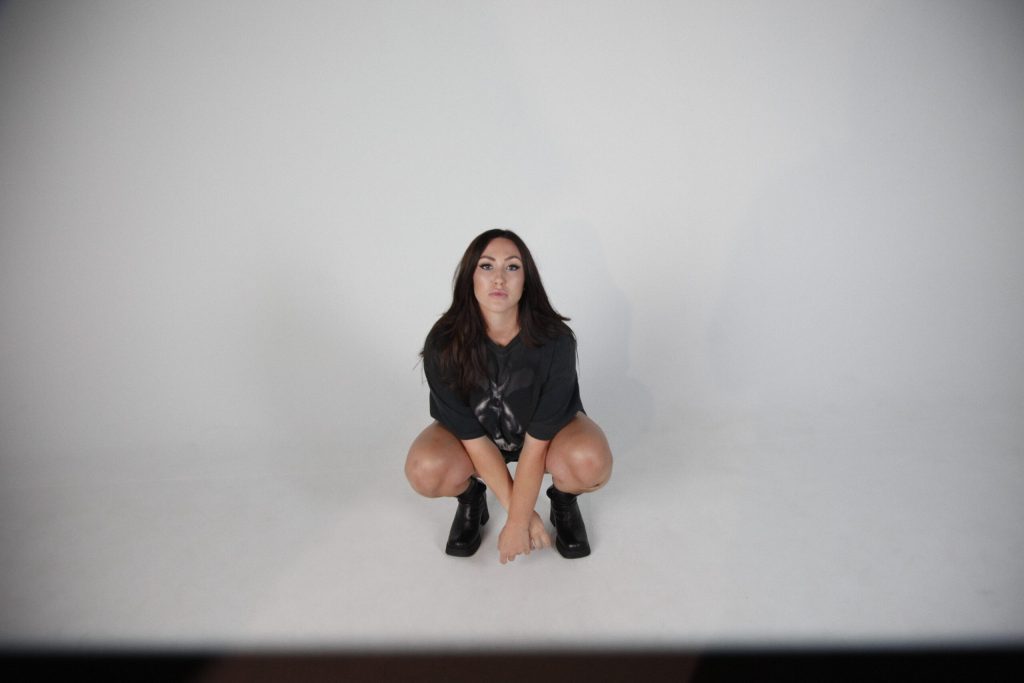 Promotional photo for "Die For Love" which sees Alyssa Reid in a white room, crouching down to the floor with her legs bare and wearing a black band t-shirt and black combat boots.