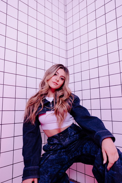 Promotional photo for "ptxd" which sees Casey Baer with blonde hair and slightly brown roots that flow past her chest, posing at the camera in a corner of a room with square patterned walls. She is wearing a fashionable denim halter top over a white crop top, paired with matching-denim trousers.