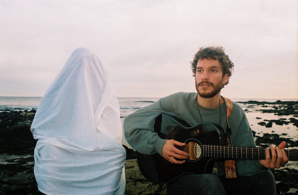 Promotional photo for "Gently, Dreamer" which sees Cayo Coco sitting down with a guitar while wearing a sea-green sweater, with the sea behind him. To his right is a person with a sheet over them like a ghost.