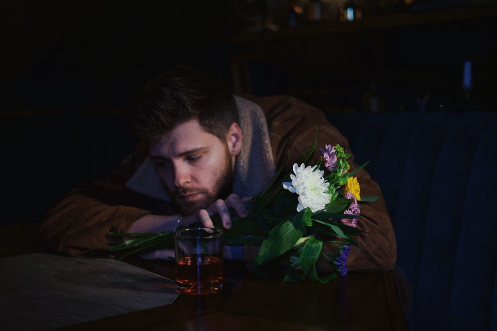 Promotional photo for "When He Was Me" which sees Charlie Rogers sitting at a table with his head on his arms which are resting on the table He has flowers in his hands and has a sad face.