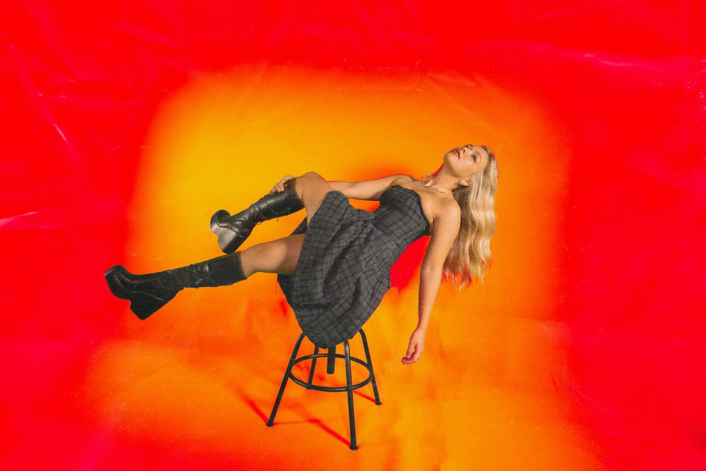 Promotional image for the music video to "Growing Pains" that sees Connie Talbot lying on a stool wearing a black dress with black knee-high boots. Her golden-blonde hair is falling down to the ground and she's balancing on the stool with her back. The background is bright orange just like the music video.