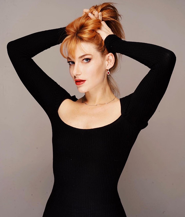 Promotional photo for "Gasoline" that sees Coyote Eyes posing for a photo shoot in front of a grey background, wearing a black square-collared long-sleeved top. She's holding her auburn hair up with both her hands.