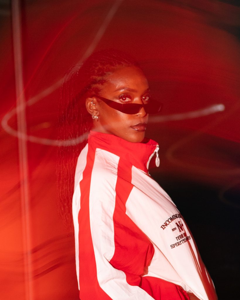 Promotional photo for "Mo Farah" which sees DEJA wearing a red and white jacket zipped to the top, with thin dark shades on. She's standing to the right with her head turned towards the camera and her long braided hair flying behind her.