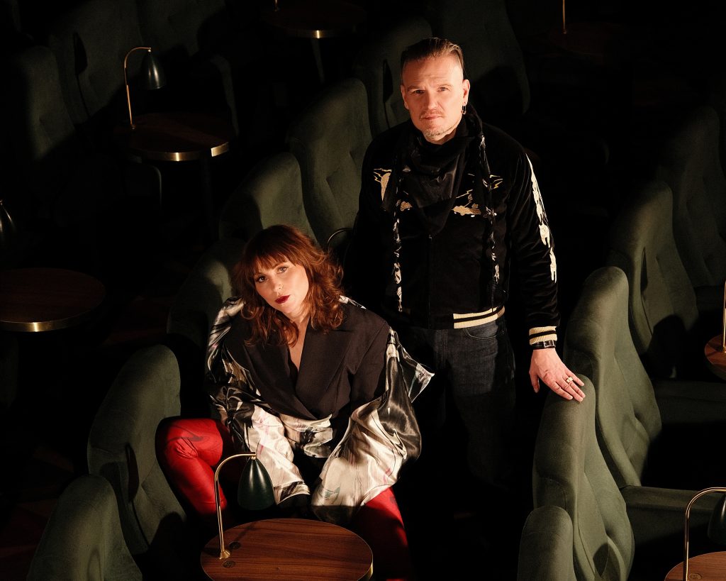Promotional photo for "Us Against The World" EP which sees the Little Lies duo in a theatre stalls posing for a photograph. Anna Maria Espinosa is sitting down, sidewards in one of the green chairs, wearing black, while Mikael Nordgren is standing behind her, also dressed in black.
