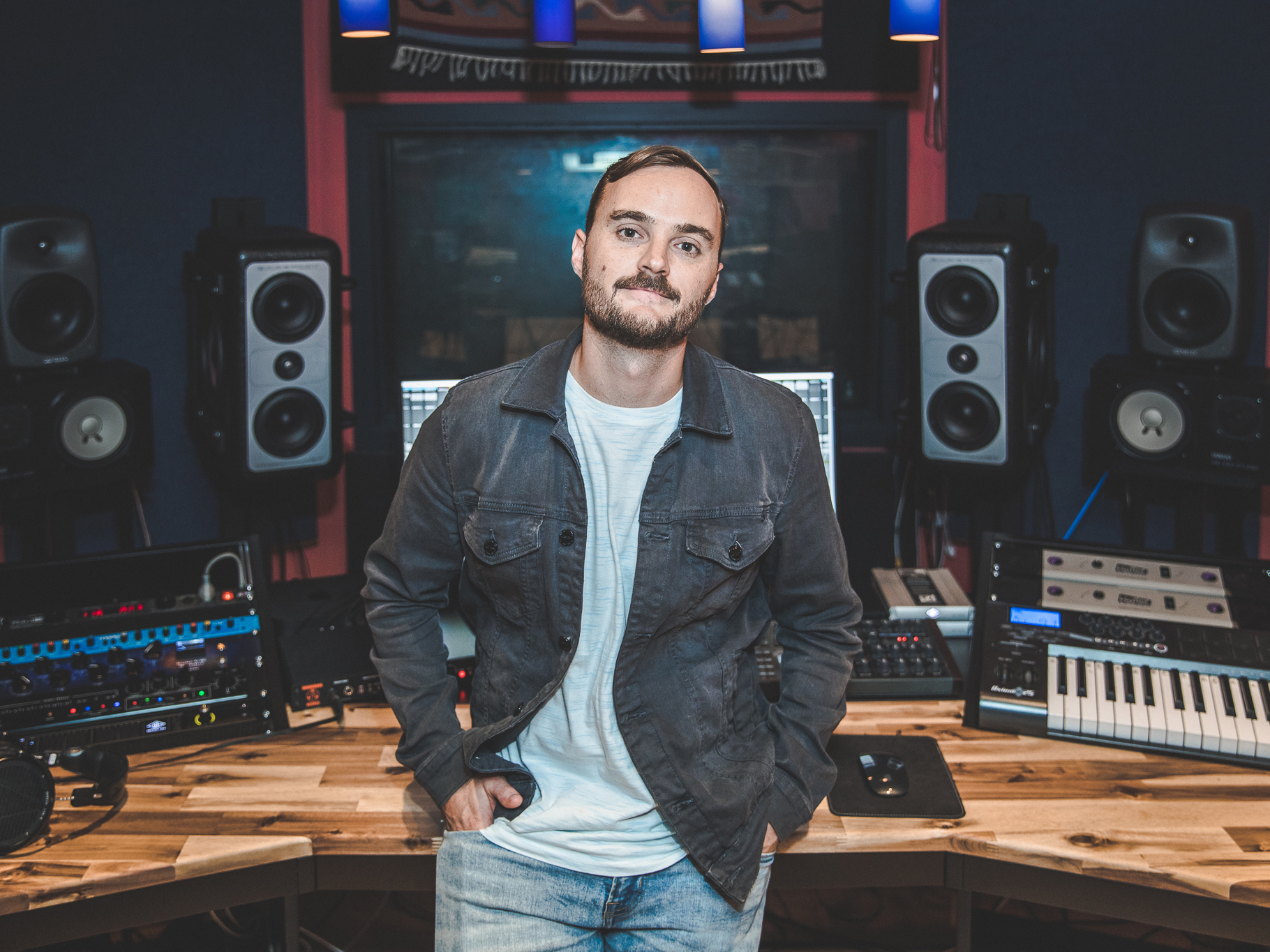 Founder and CEO of Westwood Recordings and one-half of The Funk Hunters, Nick Middleton, poses in the studio with the decks behind him. He's leaning against the desk, with his hands in his pockets, wearing a dark grey jacket over an off-white t-shirt and light blue jeans.
