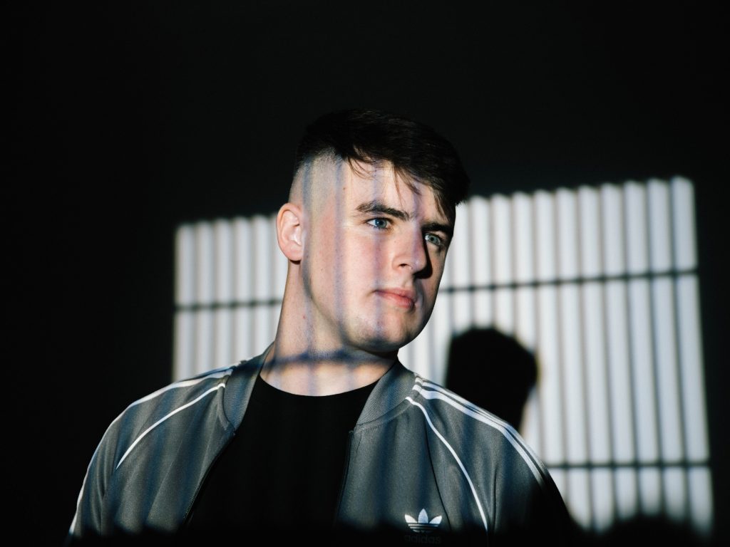 Promotional photo for "Something More" which sees Shane Codd looking to the right wearing a grey Adidas jacket over a black t-shirt. He's standing in front of a grated window and the sun is shining on him.