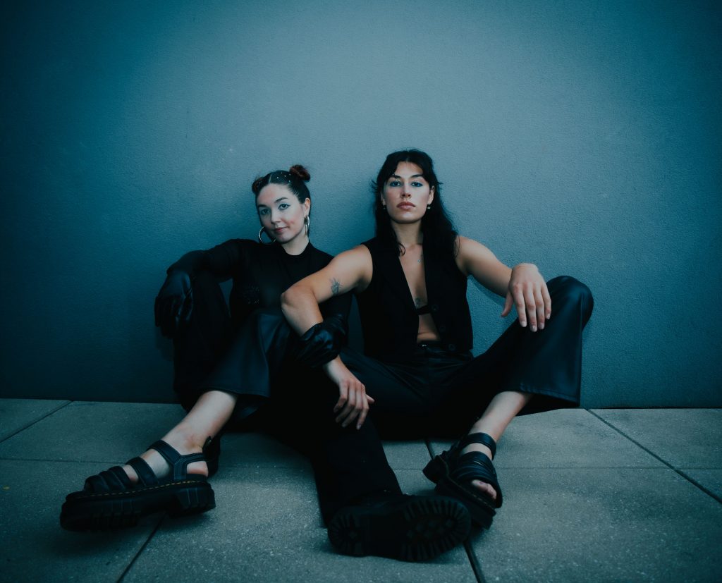 Promotional photo for "Lungs" which sees the duo FIIZ sitting on a tiled floor with their backs against a blue wall, with both women wearing black with their arms resting on their knees.
